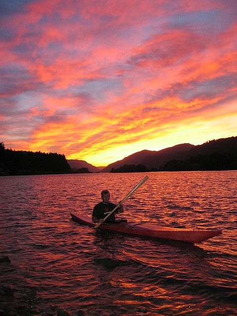 A boy paddling a kayak on calm waters during a sunset, with vibrant orange and pink hues in the sky reflecting on the water's surface, creating a serene summer scene.