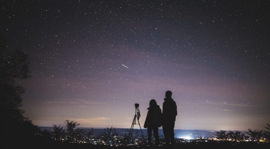 A couple standing together under a starry night sky, gazing upward with wonder and awe, surrounded by the beauty of the cosmos.