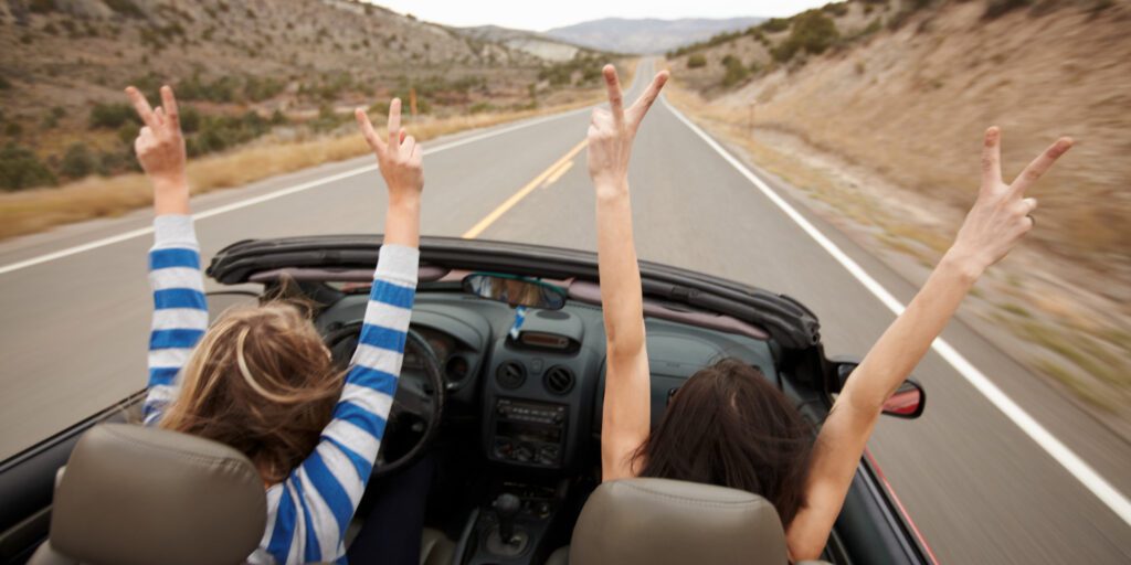 Two young women in a car during a road trip, smiling and enjoying the scenic journey with open windows and lush landscape passing by.