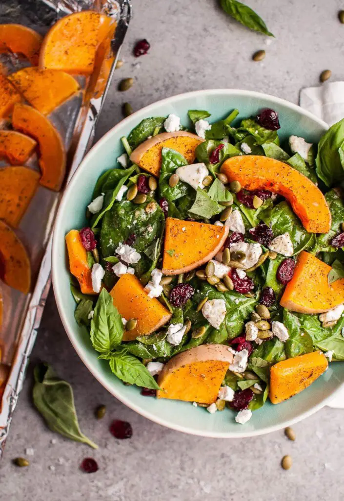 A colorful salad composed of fresh butternut squash cubes, vibrant green spinach leaves, crunchy nuts, and cranberries, drizzled with a tangy vinaigrette, served on a white plate.