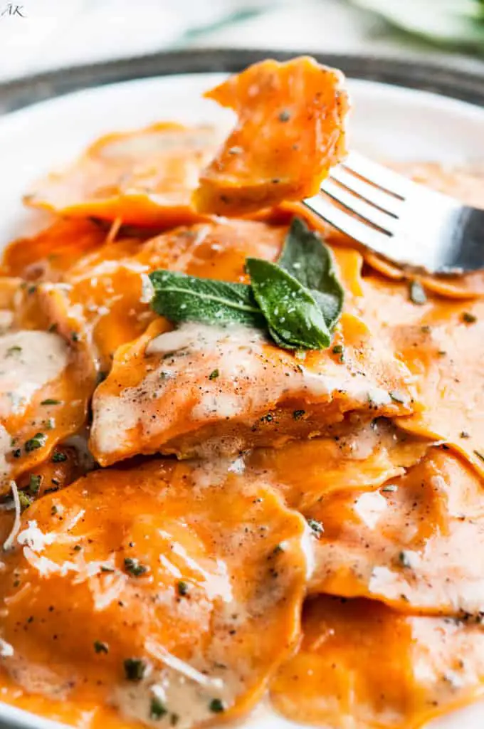 Close-up of golden brown butternut squash and sage ravioli arranged neatly on a white plate, drizzled with a rich, creamy sauce and garnished with freshly chopped herbs.