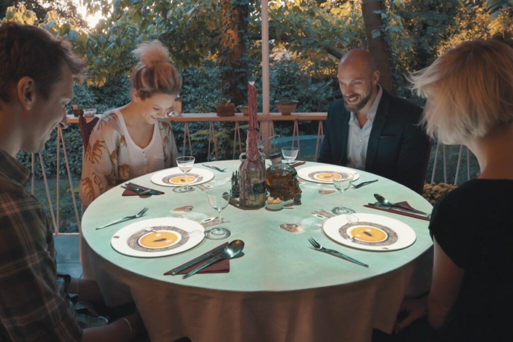 Four friends seated around a dinner table, laughing and enjoying a meal together, with plates of food and drinks in front of them, creating a warm and convivial atmosphere.