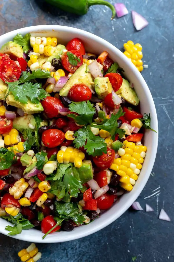 A bowl filled with vibrant corn and bean salad featuring colorful bell peppers, black beans, sweet corn kernels, and diced tomatoes, seasoned with Southwest flavors, perfect for a summer gathering.