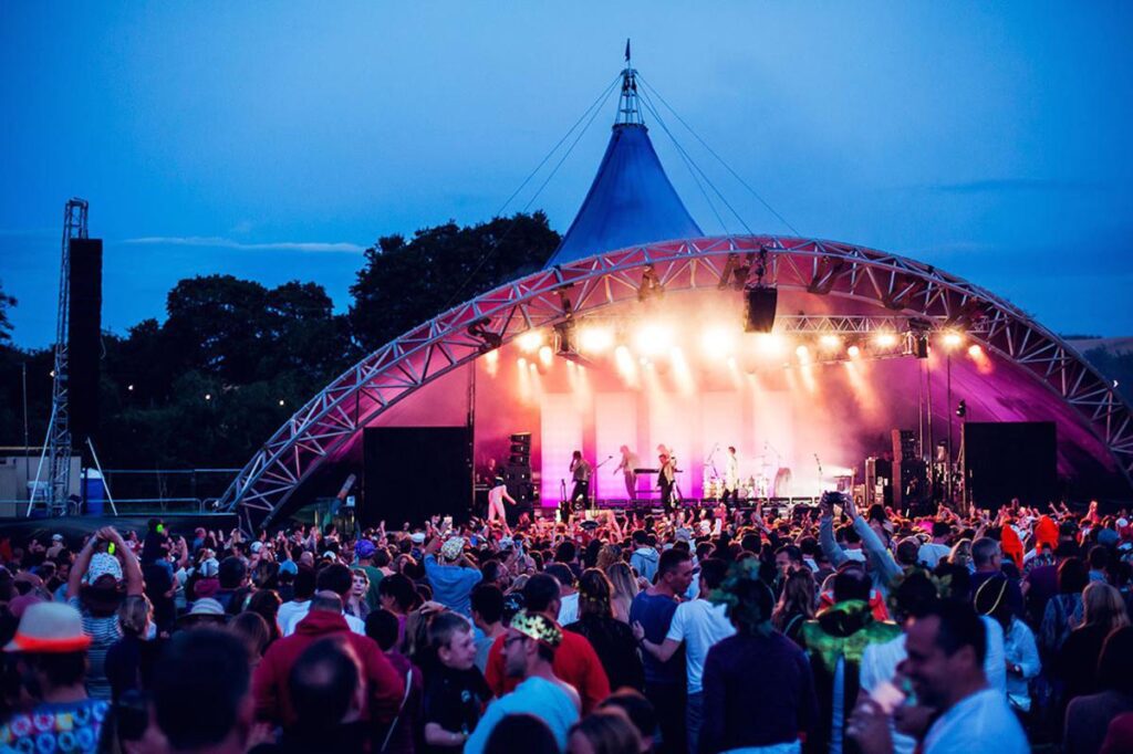 A vibrant cultural summer festival night scene with people gathered around a stage, enjoying live music performances, singing, and dancing under colorful lights and decorations.