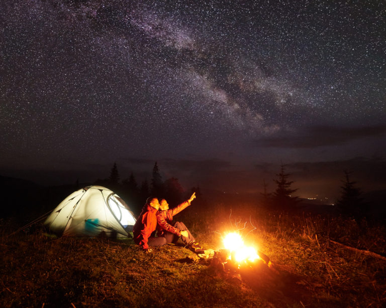 A couple sitting together beside a campfire under a starry night sky, surrounded by tents and wilderness, enjoying a romantic camping experience.